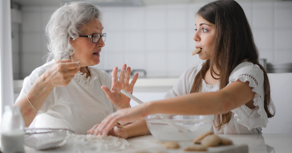 Calm senior woman and teenage girl in casual clothes looking at each other and talking while eating cookies and cooking pastry in contemporary kitchen at home