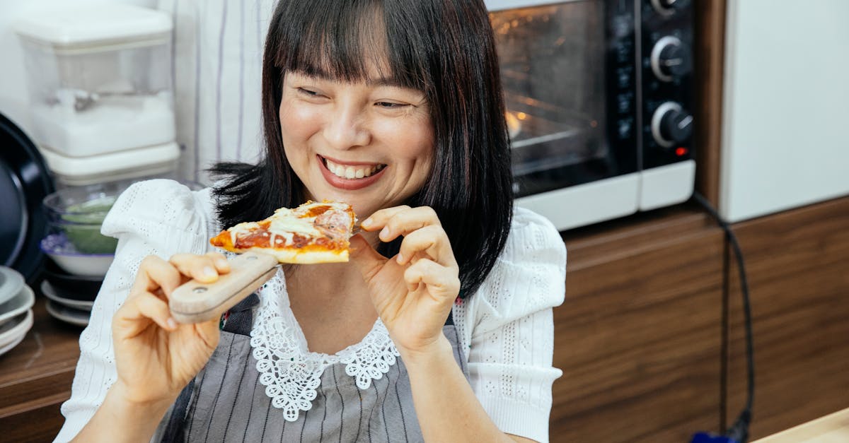 Happy Asian woman enjoying eating slice of cheesy pizza sitting at table in kitchen