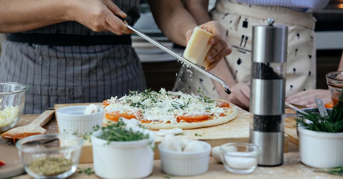 Unrecognizable female cooks grating cheese on homemade pizza at table with various ingredients and condiments in kitchen during cooking process