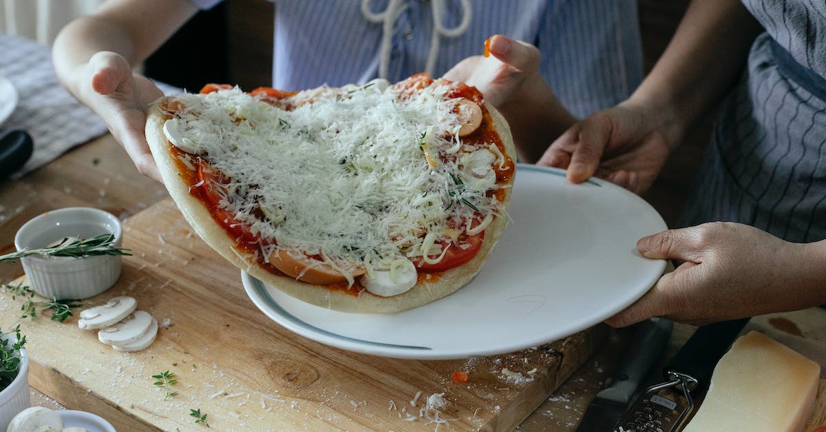 High angle of crop anonymous women transferring uncooked yummy homemade pizza on platter during preparation to baking in kitchen