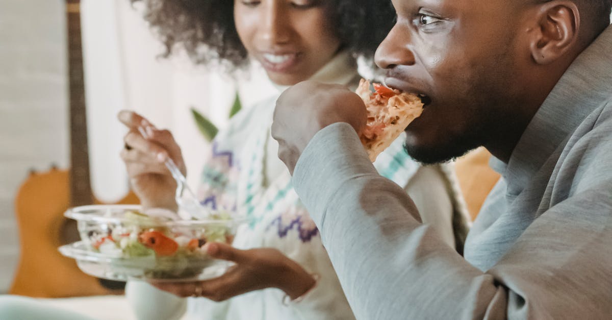 Surprised African American crop male eating pizza while ethnic girlfriend having snack with salad