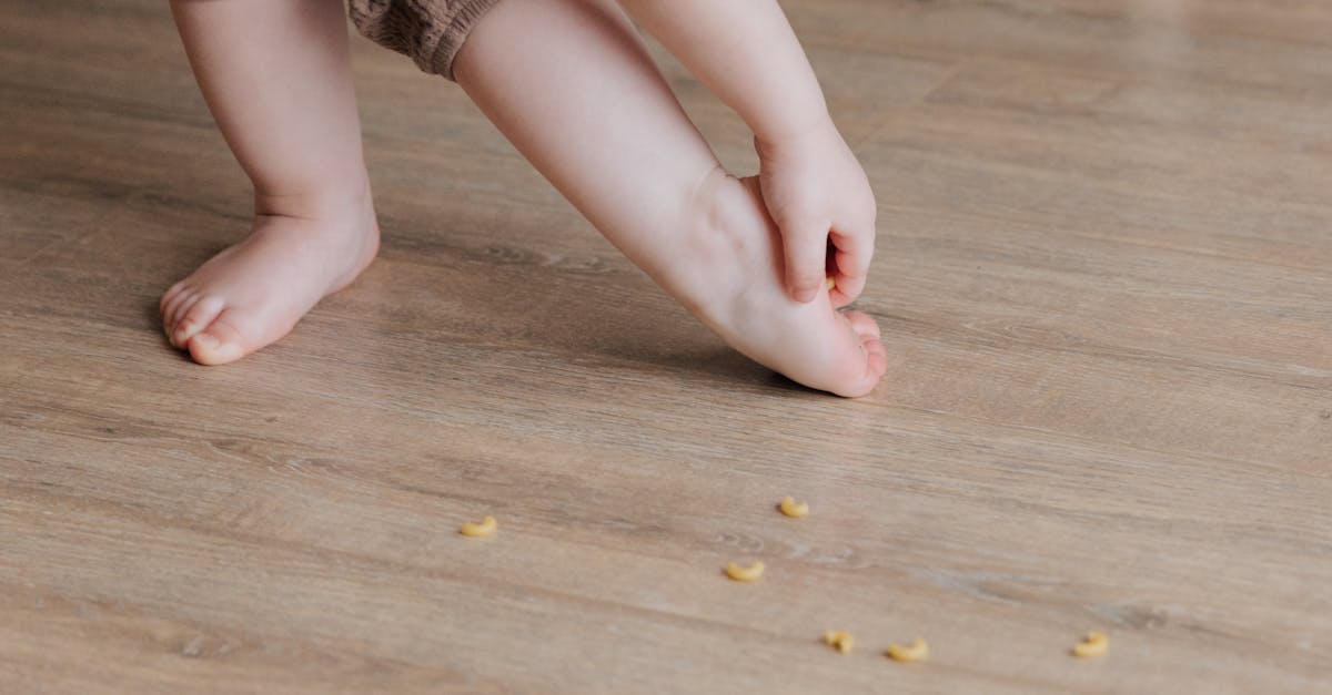 Crop faceless toddler standing barefoot on floor and trying to remove stuck pasta from foot while playing and developing fine motor skills at home