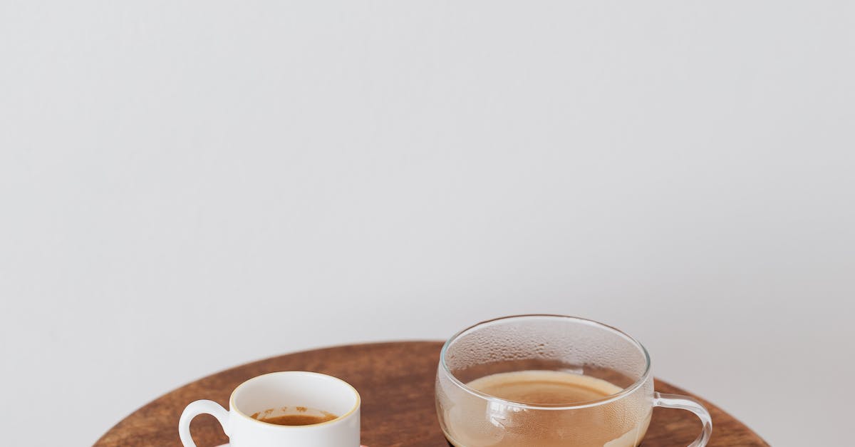 Fresh coffee in ceramic white cup and wide glass cup placed on round wooden table near white wall