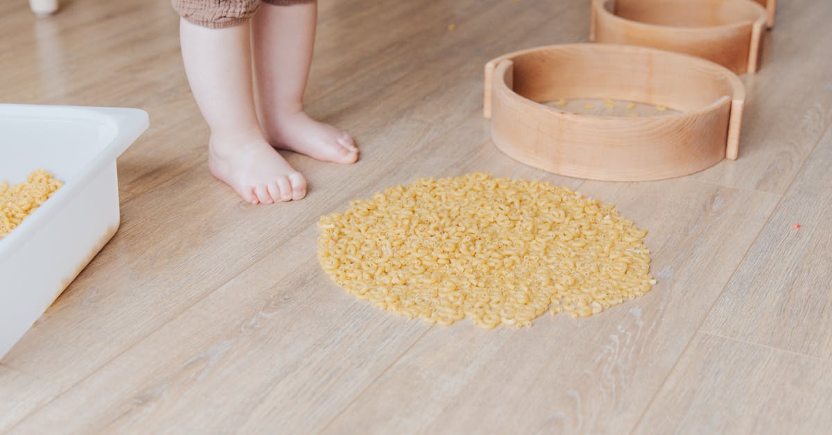 Crop faceless toddler standing barefoot on floor and playing with round wooden shapes of different size and pasta developing fine motor skills at home