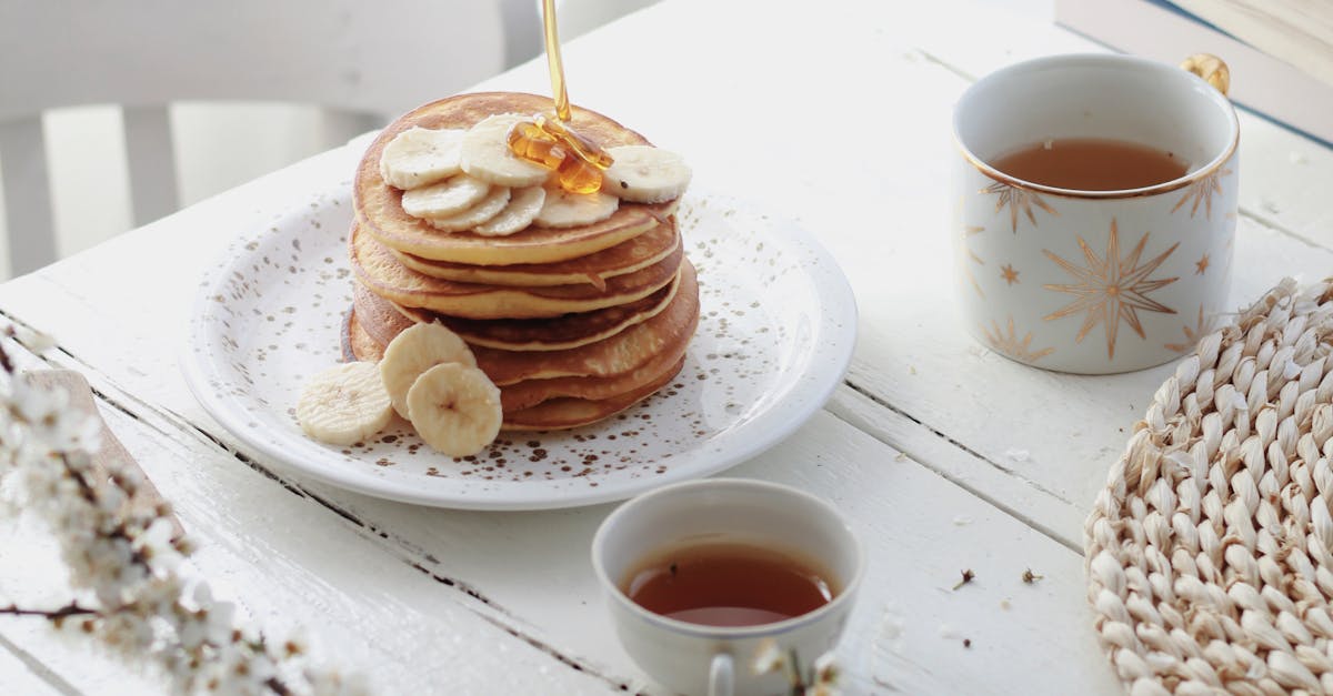 High angle of unrecognizable person adding caramel topping on tasty pancakes with banana slices served on table with cups of tea in morning