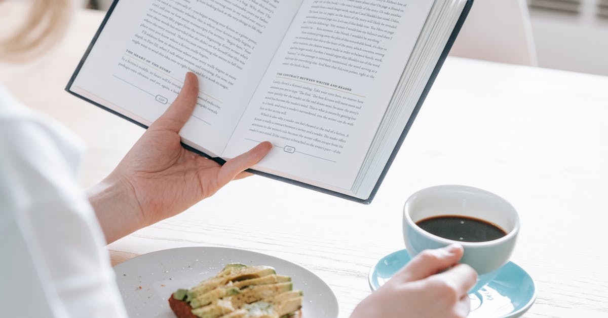 Crop unrecognizable female in white t shirt sitting at table with cup of black coffee and avocado toast while reading book