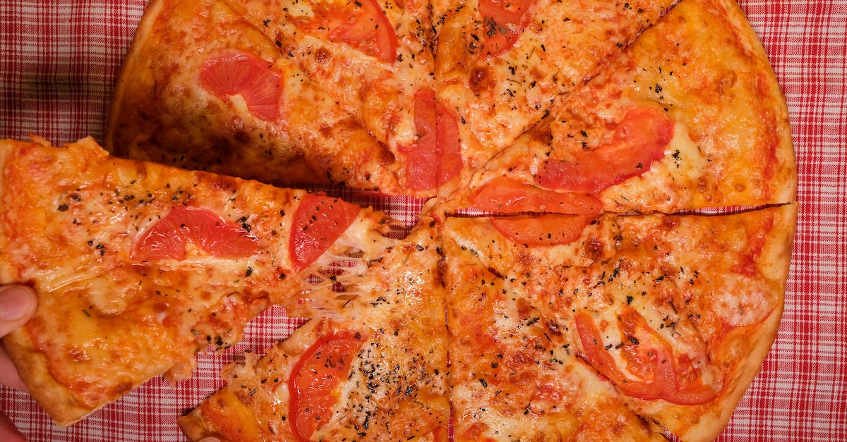 Top view of crop anonymous person taking slice of yummy pizza with tomato and oregano placed on checkered tablecloth